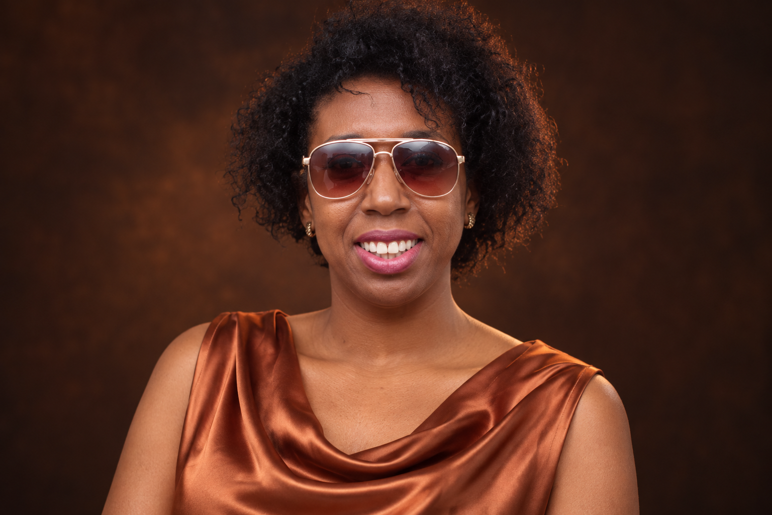 Close editorial portrait based on COCOA's reference photo, smiling in sunglasses and copper satin under warm studio light